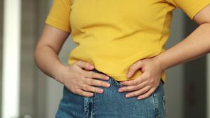 mulher com blusa amarela e calça jeans com as mãos no abdomen com cólica